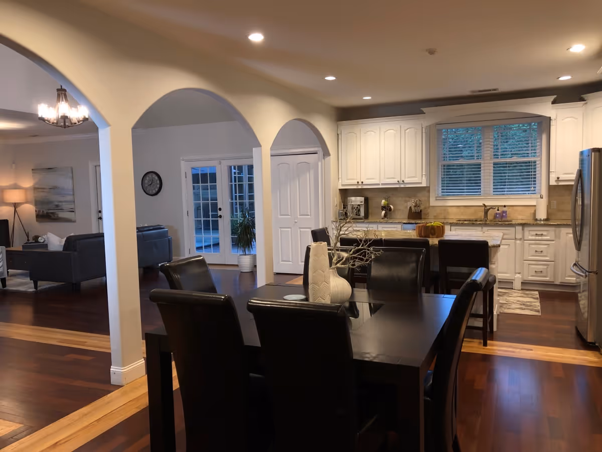 Interior view of a senior living facility showing a dining area with a dark wooden table and six black leather chairs. The dining area opens into a kitchen with white cabinets, granite countertops, a window with blinds, and stainless steel appliances. Beyond the dining area, there is a living room with dark sofas, a floor lamp, and a wall clock near French doors leading outside. The space features wooden flooring and arched doorways.