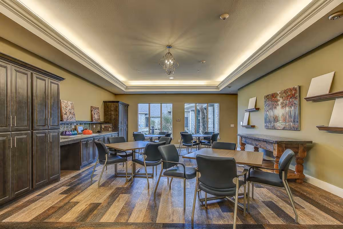 A well-lit dining room with several square wooden tables and black chairs arranged neatly. The room features wooden flooring, a decorative ceiling with recessed lighting, and large windows letting in natural light. There are cabinets and shelves along the left wall, and a wooden console table with a painting of trees on the right wall.