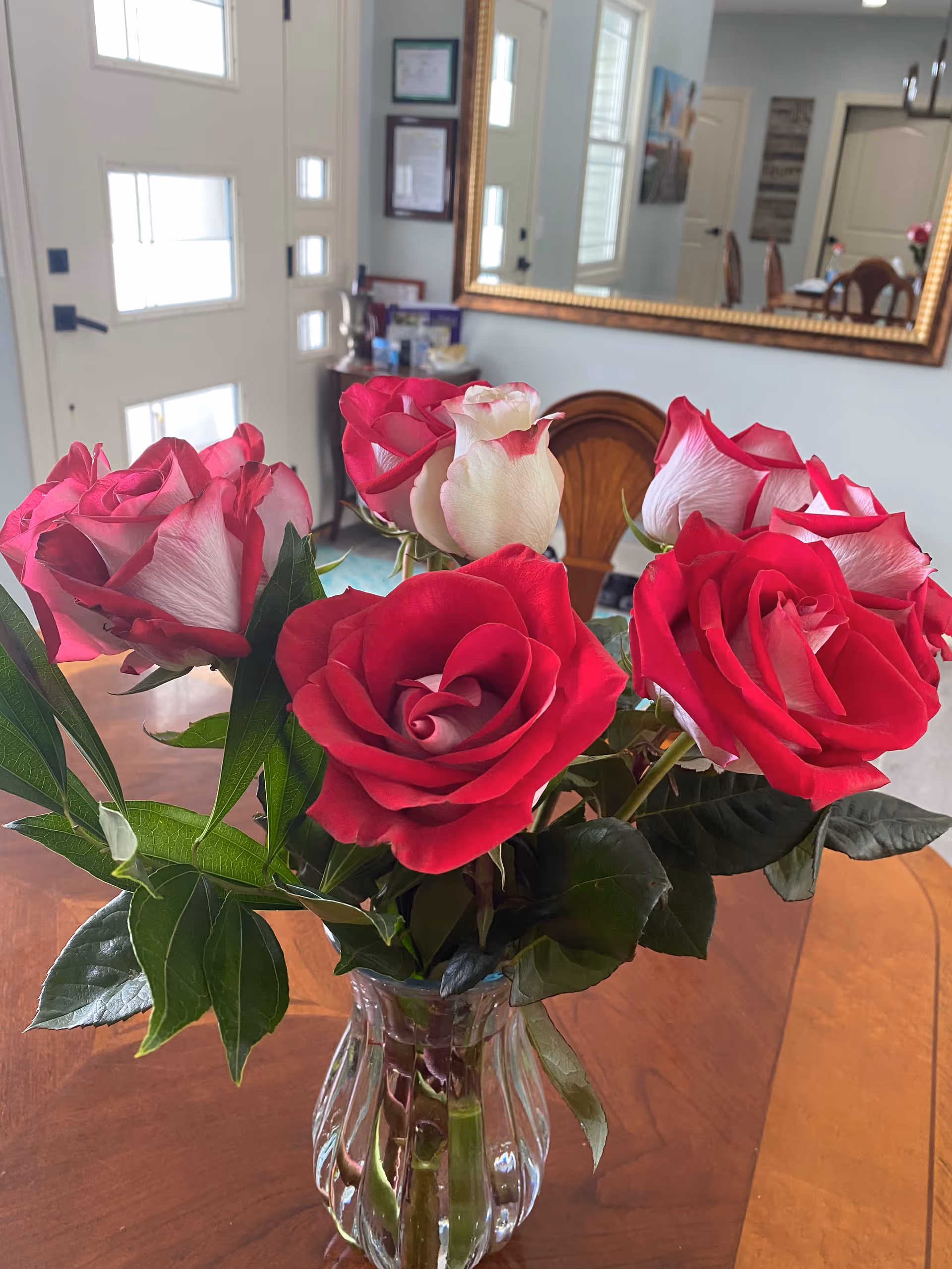 A clear glass vase filled with red and white roses sits on a wooden table in a well-lit room. In the background, there is a large mirror reflecting part of the room, including a door, framed pictures, and a dining area with chairs.