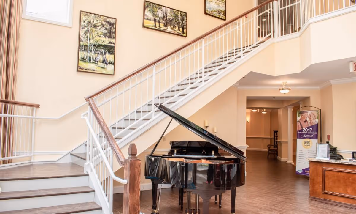 Bright interior lobby showing a black grand piano under a staircase with framed art on the walls and a nearby reception desk.