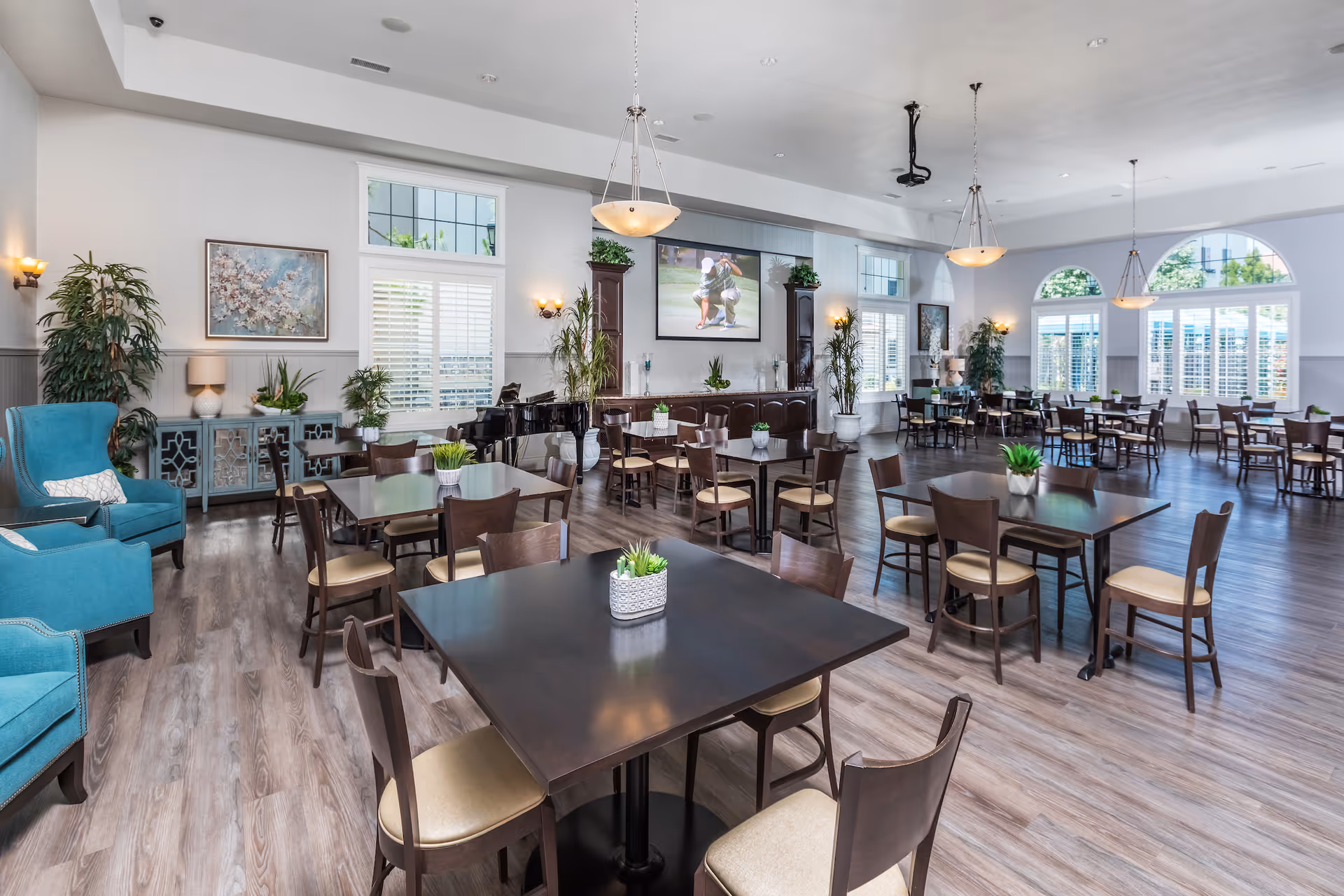 A spacious and bright dining room with multiple dark wood tables and chairs arranged neatly. The room features large windows with white shutters, allowing natural light to fill the space. There are several green plants and decorative items on the tables and around the room. Blue upholstered armchairs are placed on the left side near a sideboard with a lamp and artwork above it. A large screen is mounted on the far wall, flanked by tall wooden cabinets and more plants. The floor is wood with a light finish, and pendant lights hang from the ceiling.