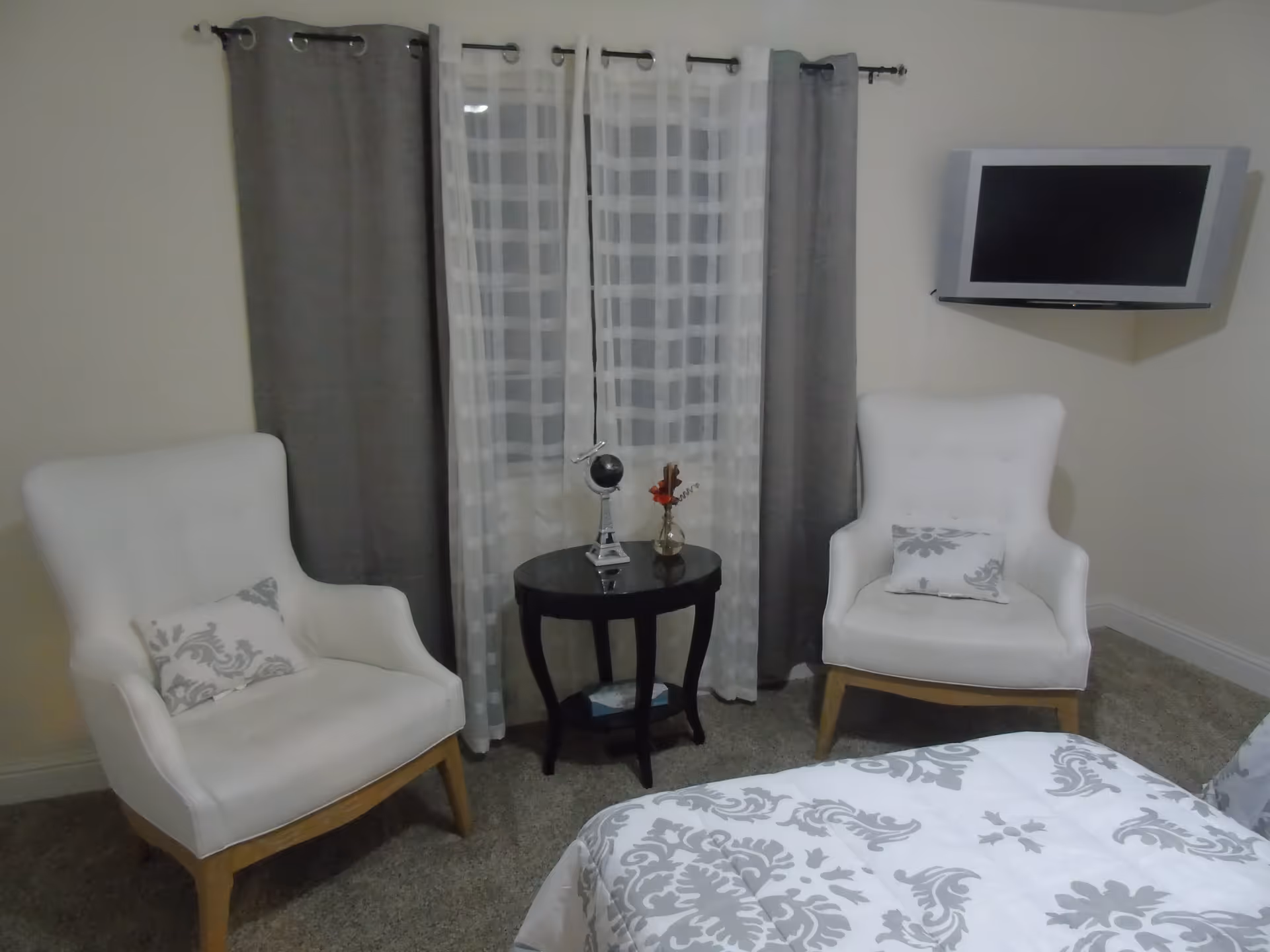 A cozy bedroom corner featuring two white armchairs with patterned cushions, a small black round table with decorative items including a small globe and a vase with flowers, a window with sheer and gray curtains, and a wall-mounted flat-screen TV.