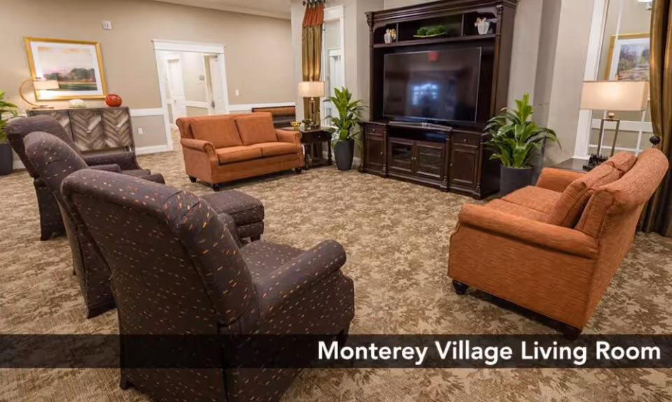 A cozy living room with two orange sofas and two patterned armchairs arranged around a large dark wood entertainment center with a flat-screen TV. The room has beige walls, carpeted floor, potted plants, lamps, and framed artwork on the walls.