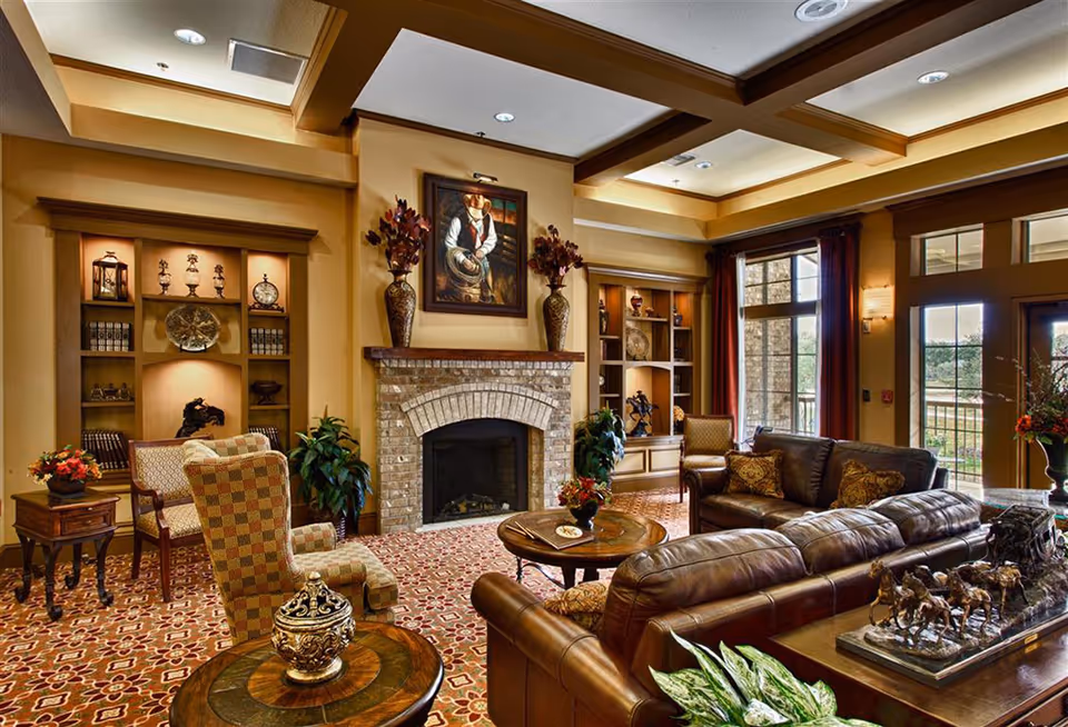 A cozy and elegant living room with a brick fireplace centered on the far wall, flanked by built-in shelves displaying decorative items. The room features a patterned carpet, leather sofas, upholstered armchairs, wooden tables with decorative vases and plants, and large windows with red curtains letting in natural light.