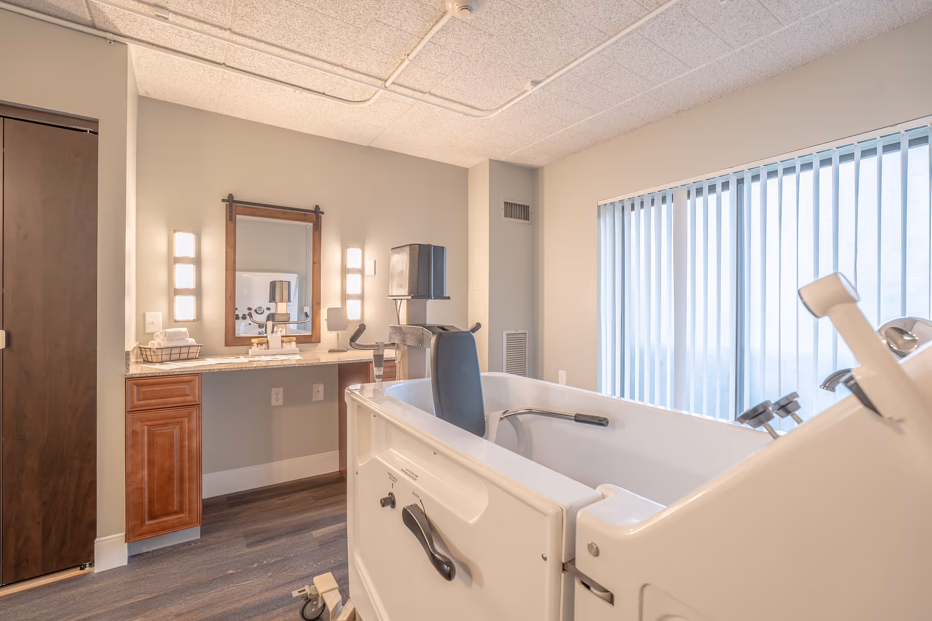 A bright and clean bathroom featuring a large walk-in bathtub with handrails and a seat. The room has a vanity with a granite countertop, a wooden framed mirror, and wall-mounted lights on either side. Vertical blinds cover a large window, allowing natural light to fill the space. The floor is covered with wood-like flooring, and there is a wooden door to the left.