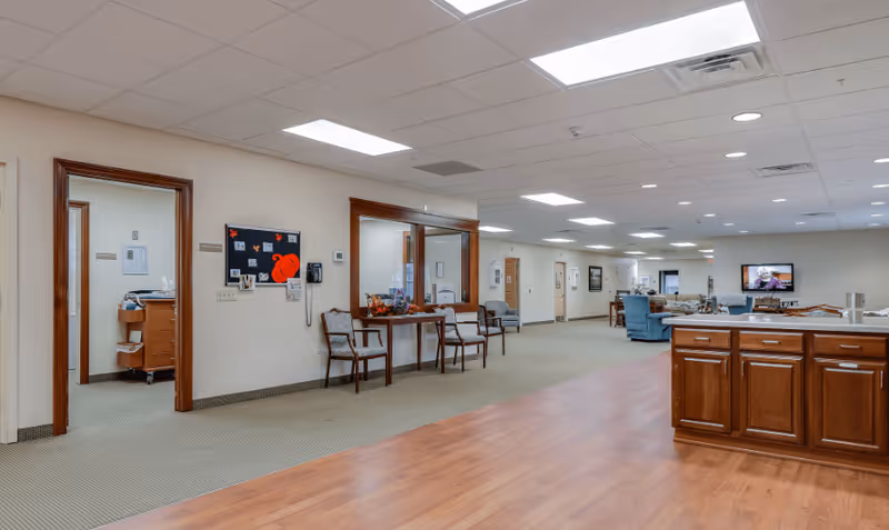 Interior view of a senior living facility hallway with a wooden cabinet on the right, chairs along the wall, and a seating area with armchairs and a television in the background. The space is well-lit with ceiling lights and has a combination of carpet and wood flooring.
