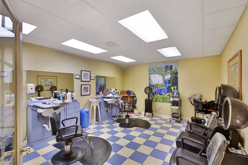 Interior of a hair salon with blue and white checkered floor tiles, several black salon chairs, hair dryers, and a counter with various hair care products. The walls are light yellow with framed pictures and a large colorful painting. The room is brightly lit with ceiling lights.