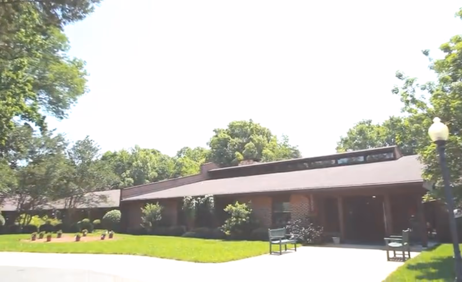 Exterior view of a single-story brick building with a sloped roof surrounded by green trees and a well-maintained lawn. There are benches and a lamppost near the entrance pathway.