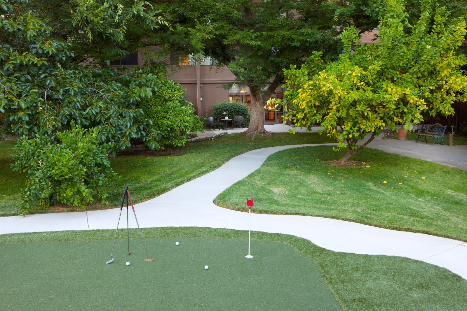 A well-maintained outdoor putting green with three golf balls and two golf clubs resting on it. Surrounding the green is a curved concrete pathway, lush green grass, and various trees and bushes. In the background, there is a building with windows and a small patio area with chairs and a table.