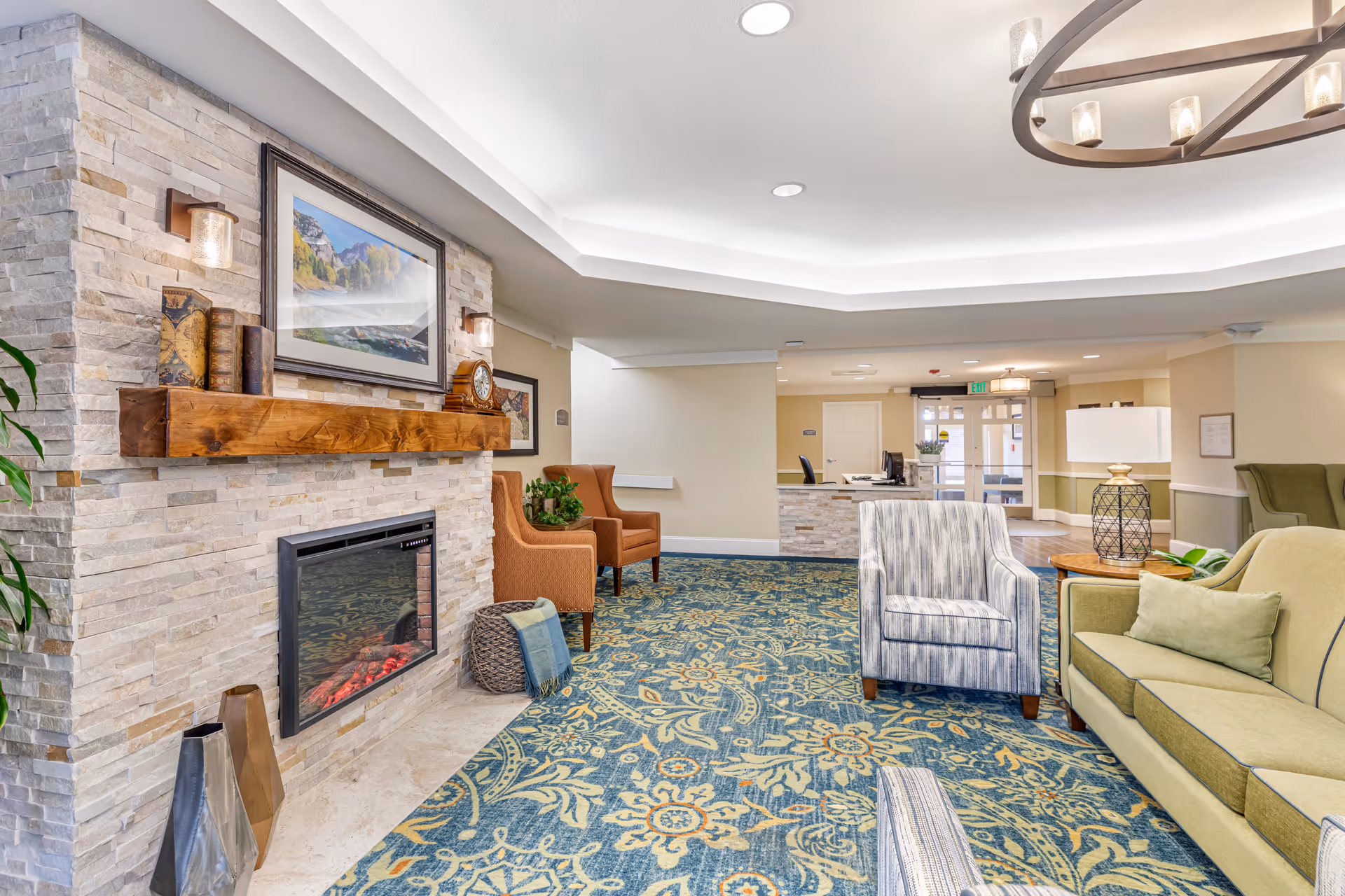 A cozy senior living facility common area with a stone fireplace, wooden mantel decorated with books and a clock, and a framed landscape picture above it. The room features patterned blue and green carpet, several upholstered chairs and a sofa, a side table with a lamp, and a reception desk in the background near the entrance.