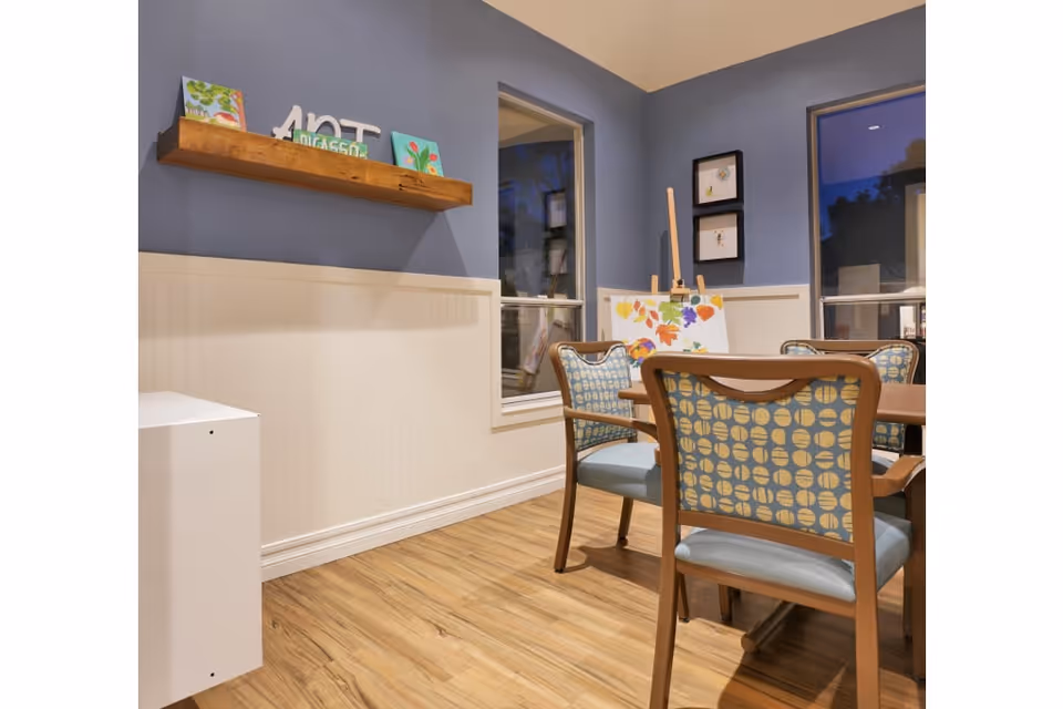 A cozy room with wooden flooring and blue walls featuring a wooden table surrounded by four chairs with patterned blue upholstery. On the wall, there is a wooden shelf holding small art pieces and a sign that says 'ART'. An easel with a colorful painting is positioned near two windows showing a nighttime outdoor view.