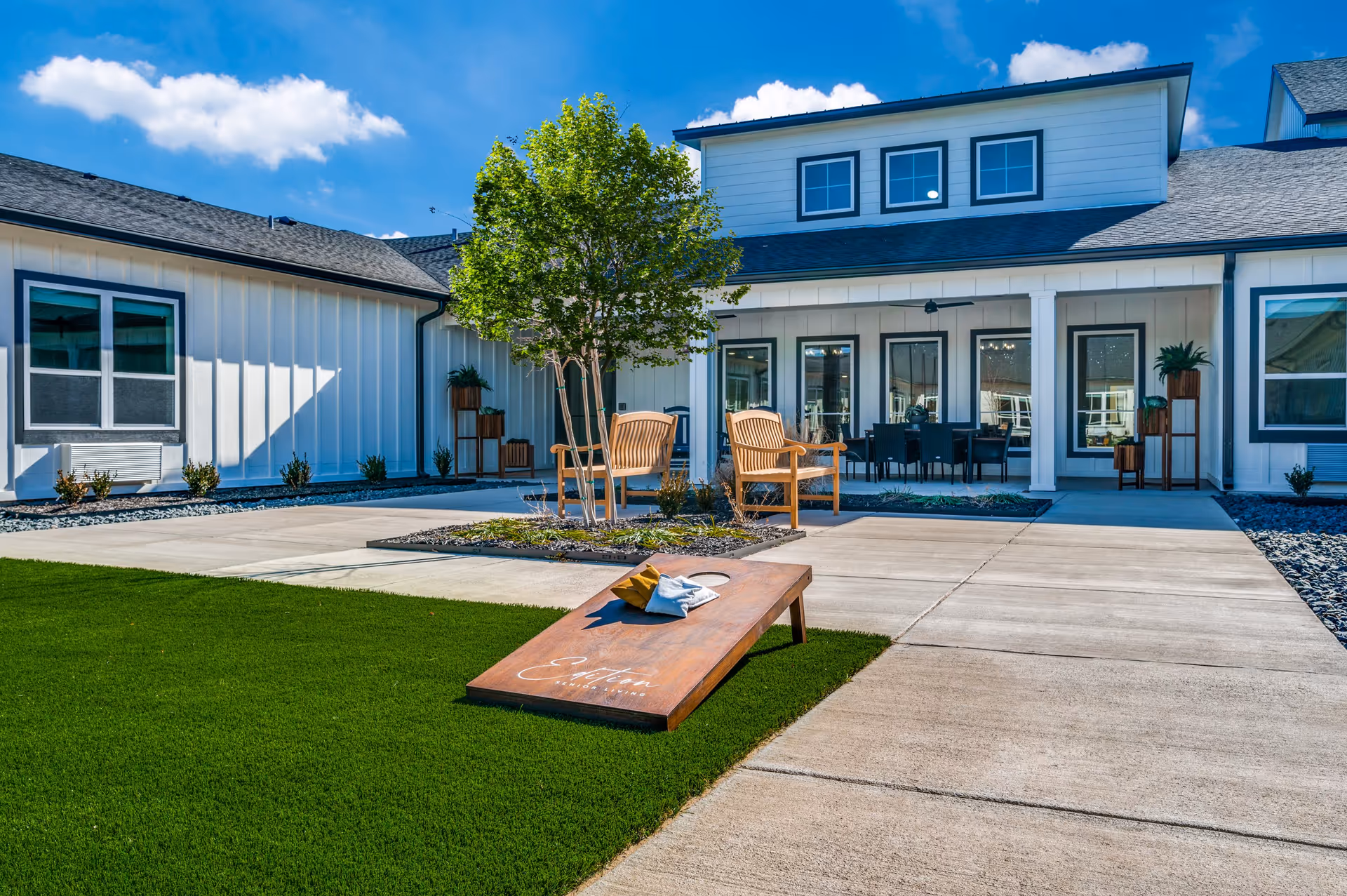 Courtyard with a cornhole board on green turf, wooden chairs and a small tree in front of a white building's covered patio.