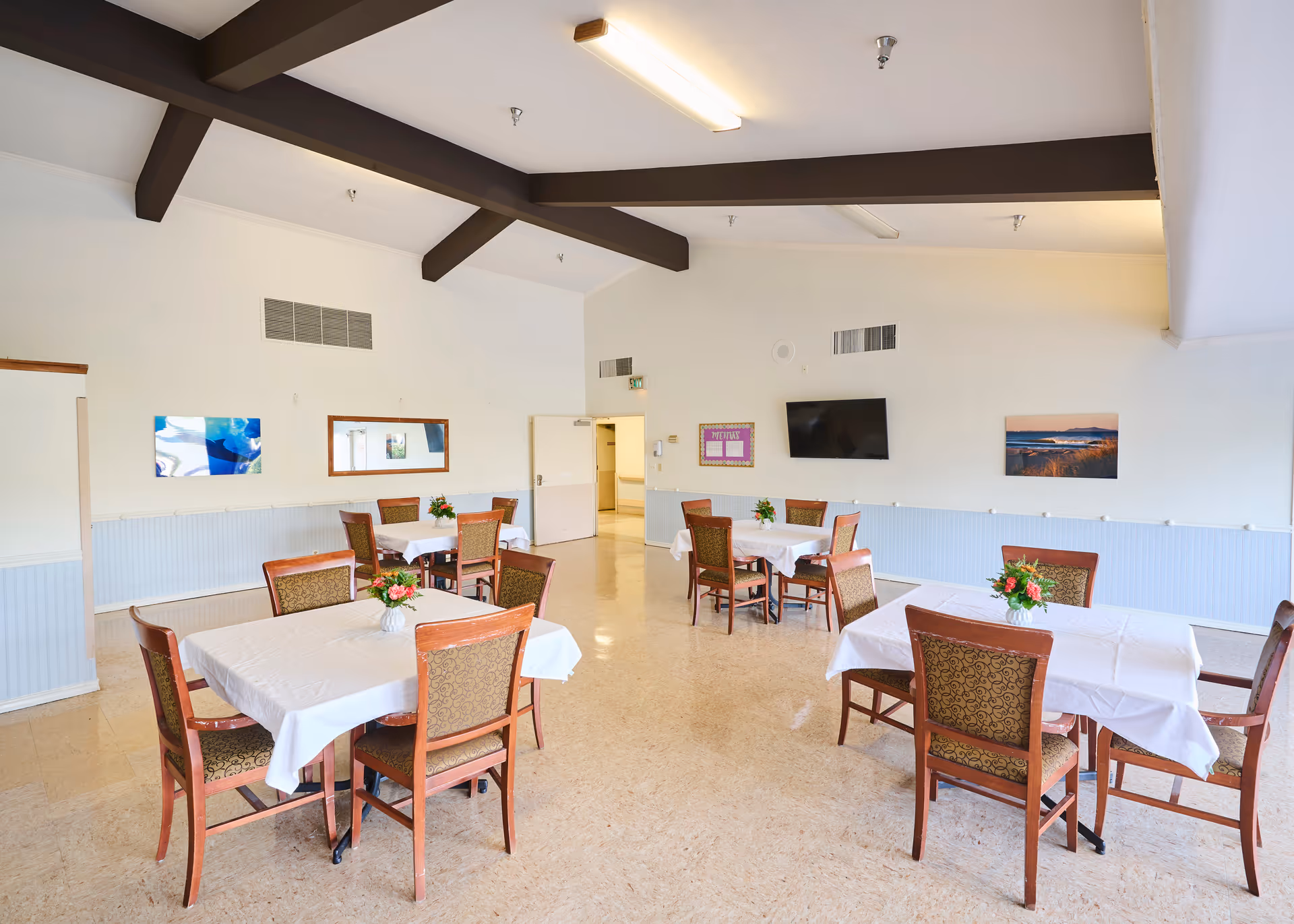 Bright communal dining room with several tables covered in white tablecloths and floral centerpieces, wooden chairs, a wall-mounted TV, and exposed ceiling beams.