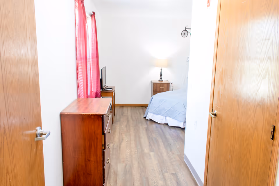 View into a small bedroom from the doorway, showing a wooden dresser, red curtains, a bed with gray bedding and a bedside table with a lamp.