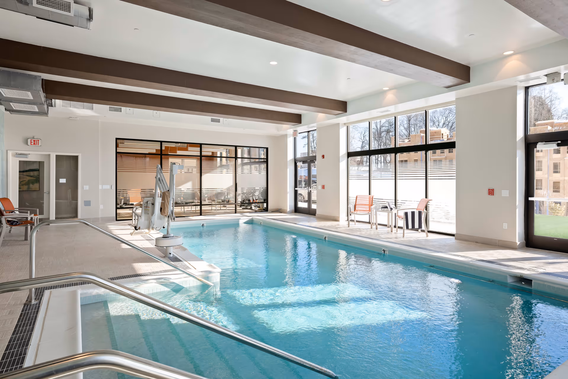 Indoor swimming pool area with clear blue water, metal handrails, a pool lift for accessibility, and seating with chairs and a small table near large windows letting in natural light.