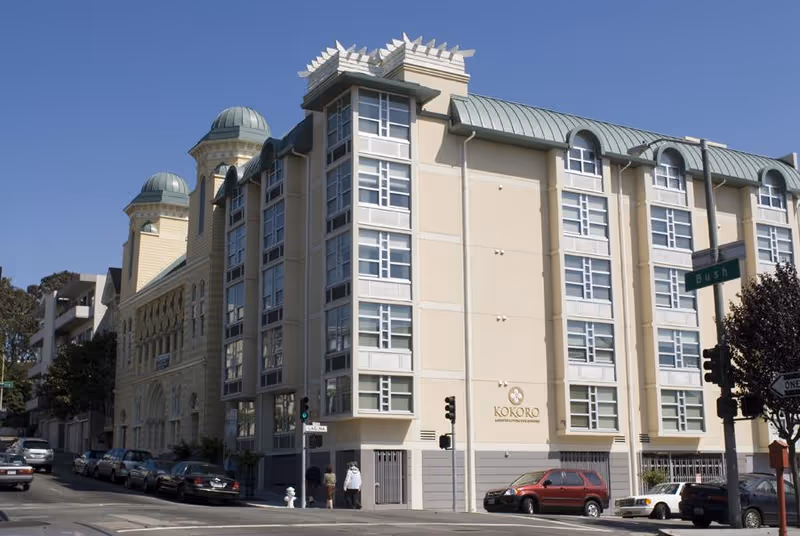 Exterior view of a multi-story assisted living facility named Kokoro Assisted Living Inc, located on a street corner with several parked cars and pedestrians nearby under a clear blue sky.