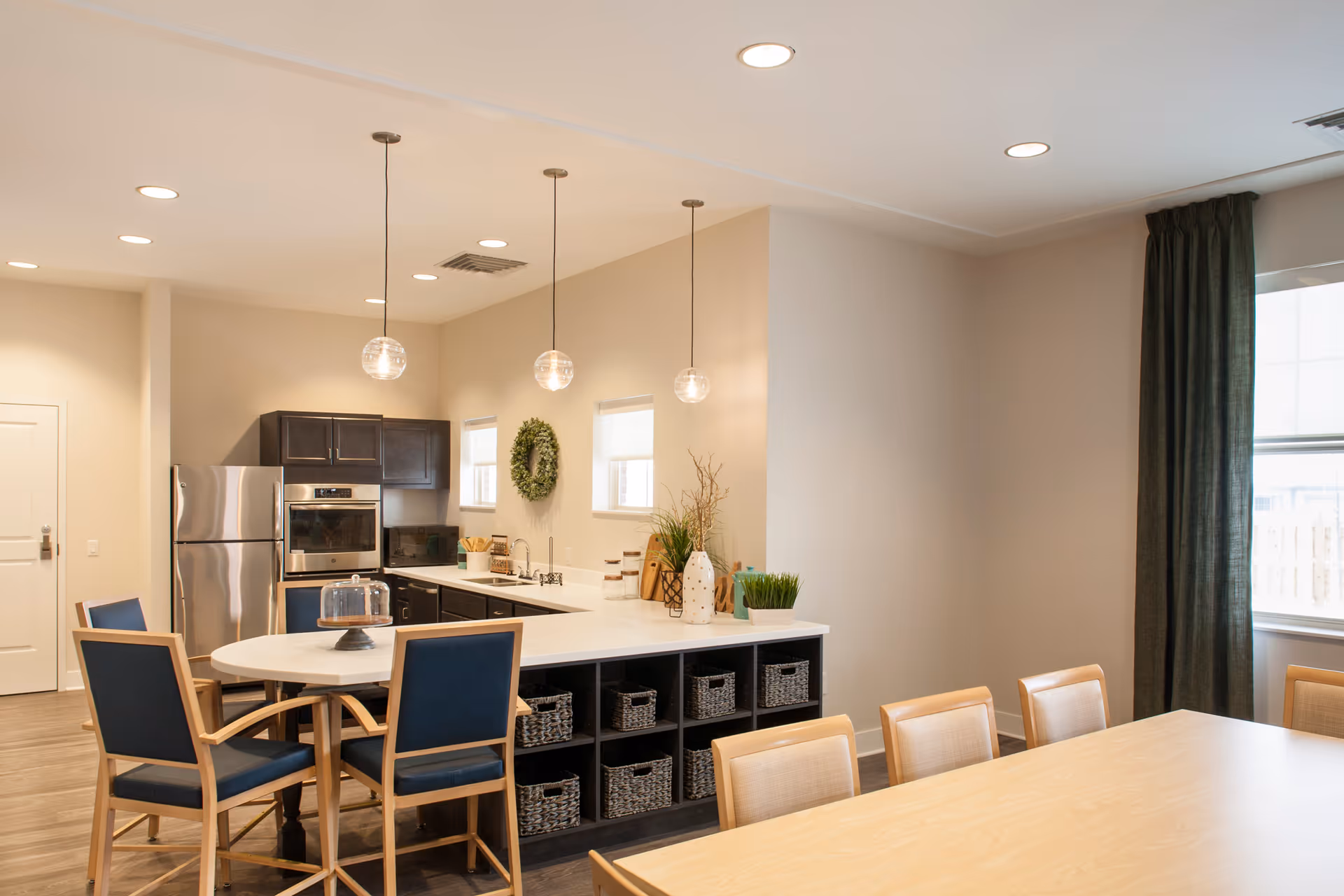 A modern kitchen and dining area with a round table surrounded by four chairs with blue cushions, a kitchen island with storage baskets underneath, stainless steel refrigerator and oven, pendant lights hanging from the ceiling, and a window with dark curtains.