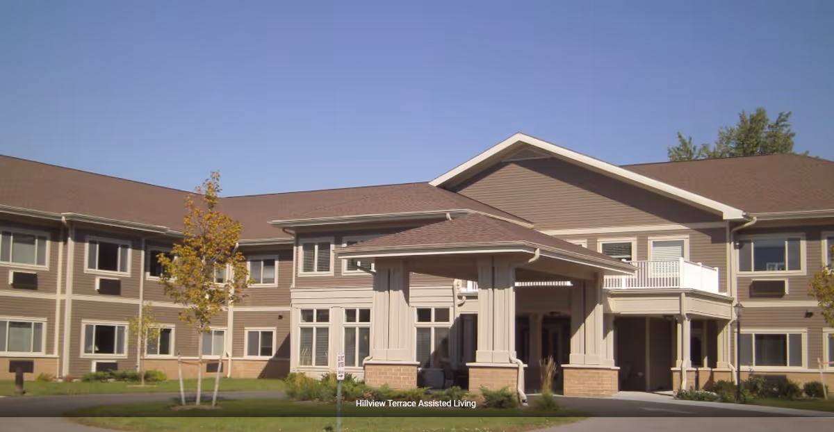 Exterior view of Hillview Terrace Assisted Living facility showing a two-story building with beige siding, multiple windows, a covered entrance with columns, and a clear blue sky.