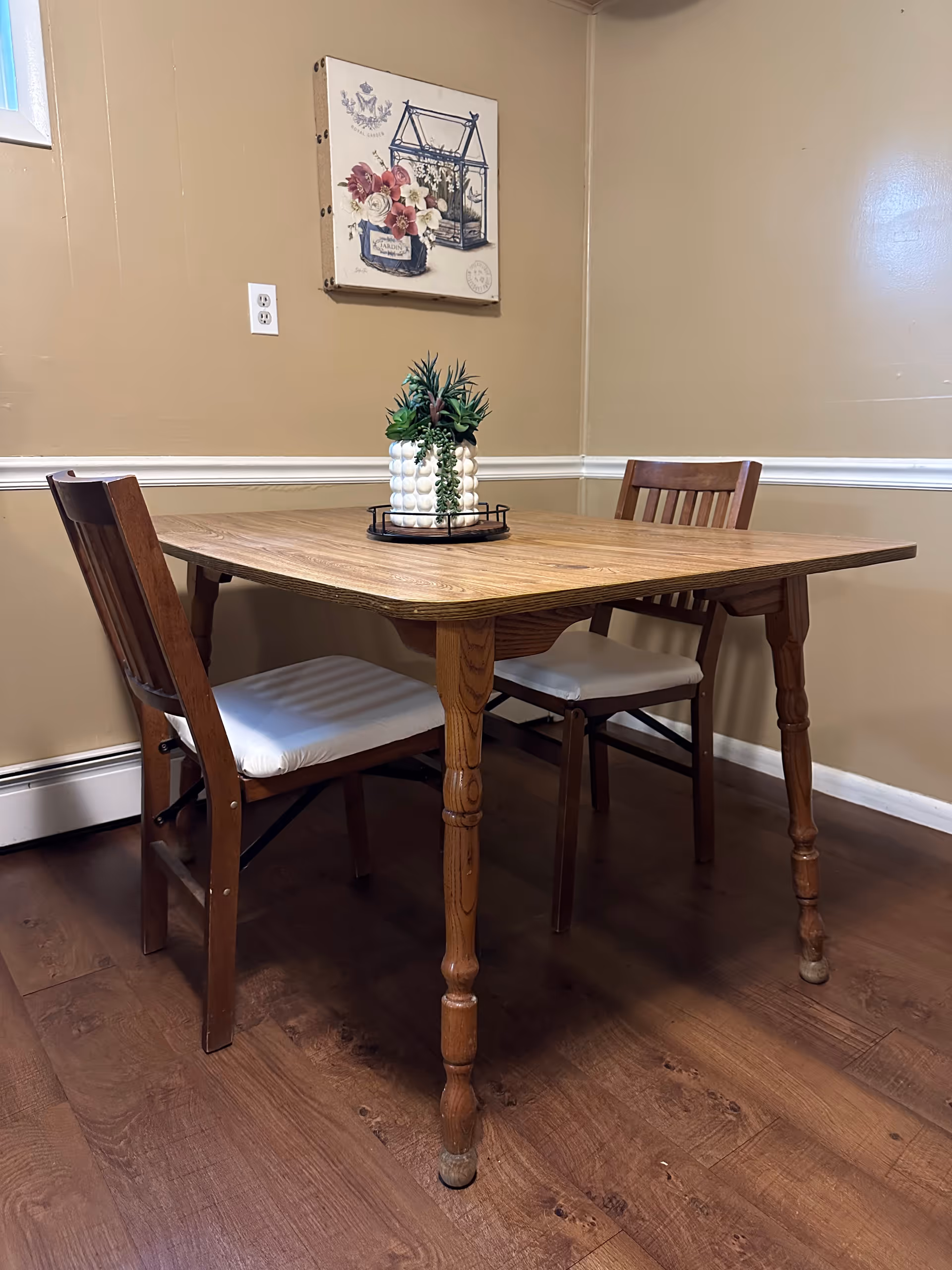 A small wooden dining table with two wooden chairs that have white cushions. On the table is a decorative white vase with green succulent plants. The room has beige walls with white trim and a framed floral artwork hanging on the wall.