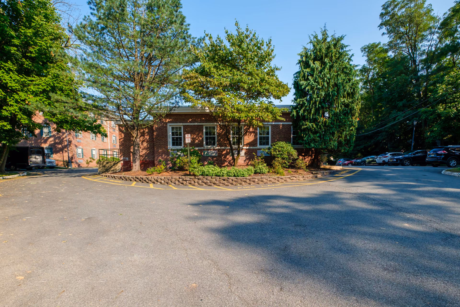 Brick building facade surrounded by trees with a paved driveway and parked cars in front.