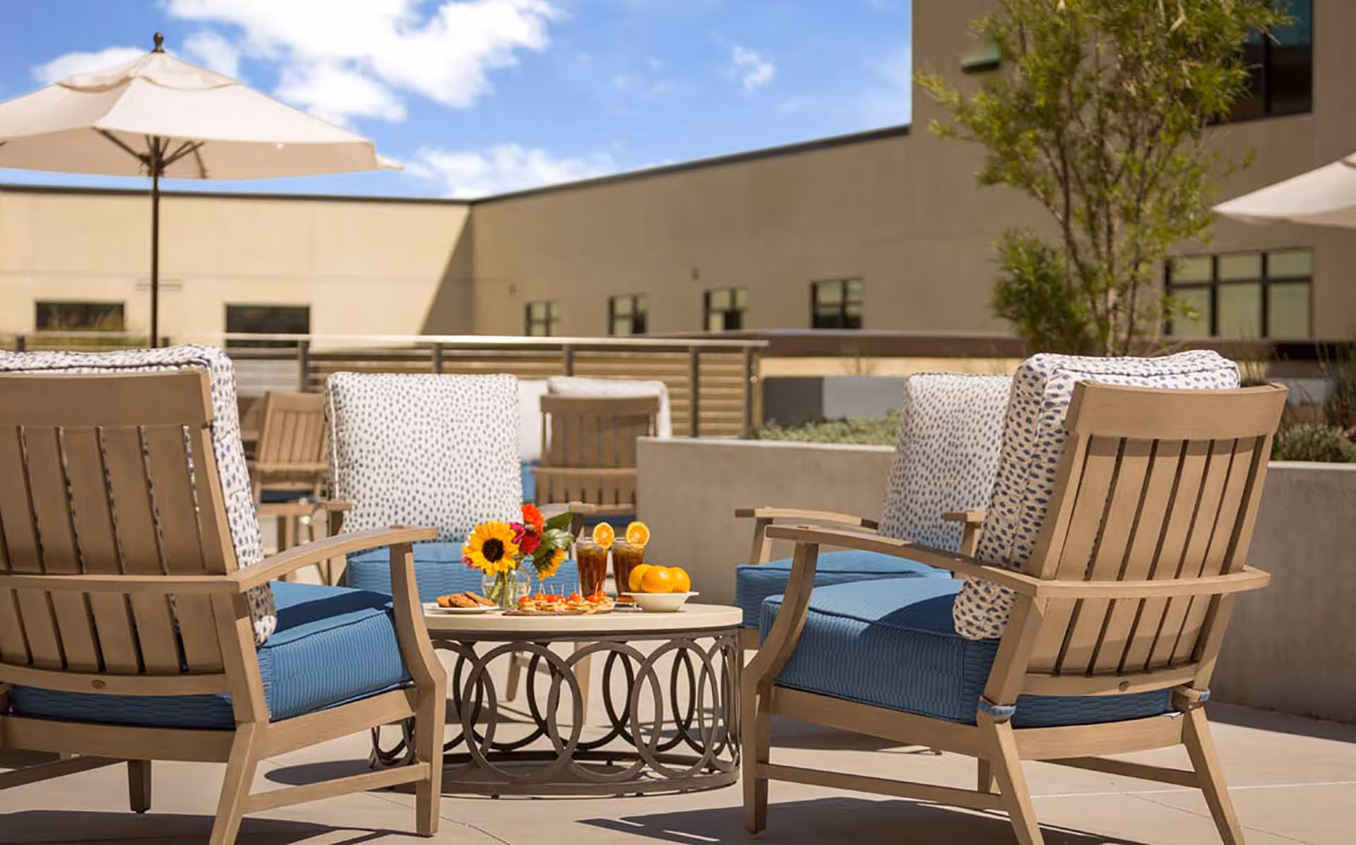 Outdoor patio area with cushioned wooden chairs arranged around a circular table holding drinks, snacks, and a small flower vase, with umbrellas and a building in the background under a partly cloudy sky.