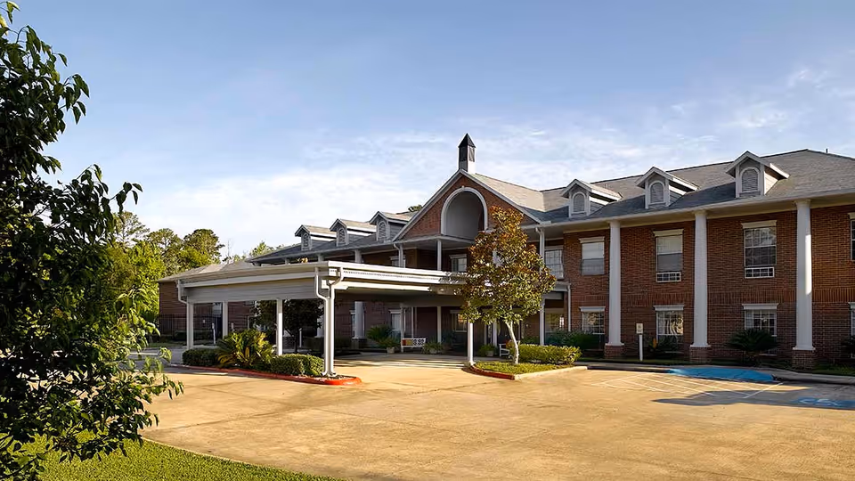 Exterior view of a two-story brick building with white columns and a covered entrance, surrounded by greenery and a parking area under a clear sky.