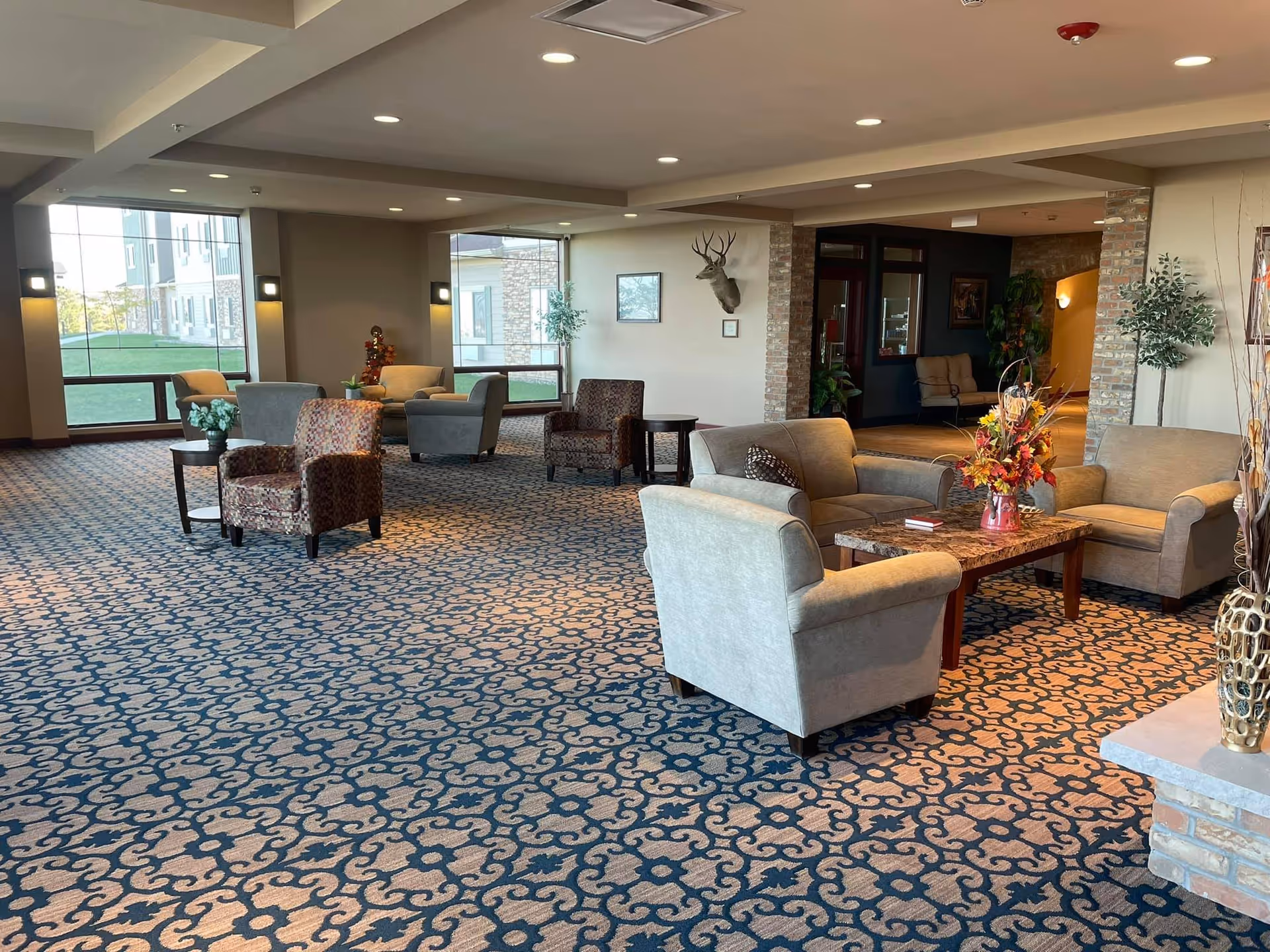 A spacious and well-lit common area in an assisted living facility featuring patterned carpet, multiple seating arrangements with armchairs and sofas, a coffee table with a floral arrangement, large windows letting in natural light, and decorative elements including a mounted deer head and plants.