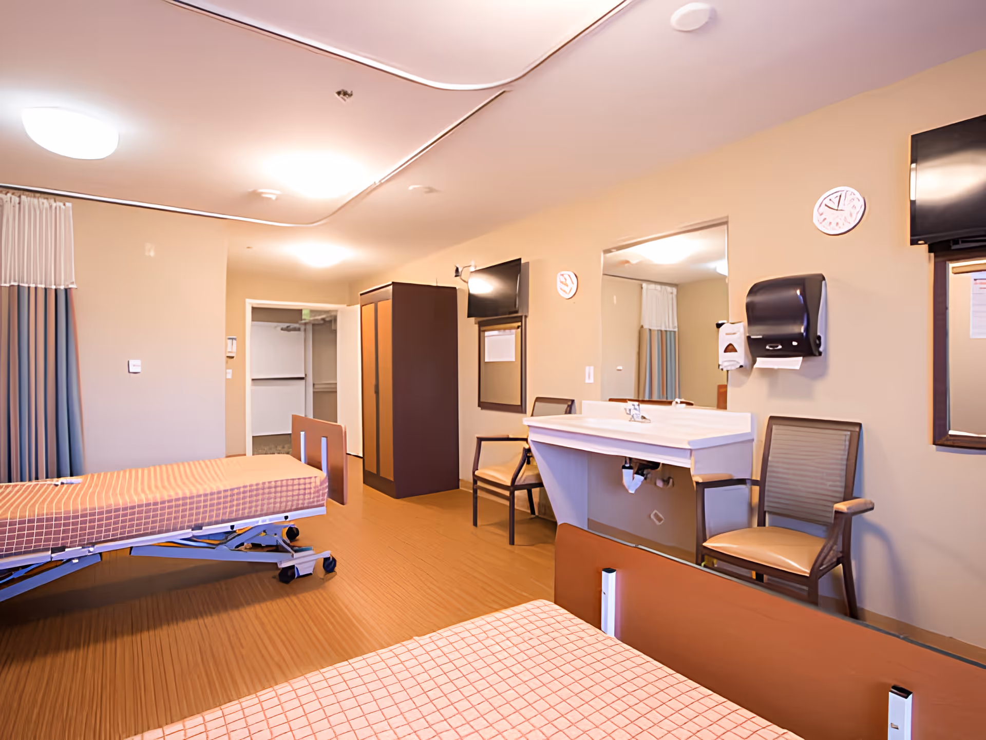 A bright assisted-living bedroom with two adjustable beds, a sink and chairs, a wall-mounted TV, and a wardrobe.