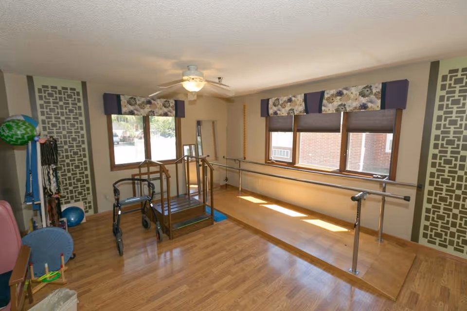 A rehabilitation therapy room with wooden flooring, parallel bars for walking practice, a small set of stairs with handrails, a walker, and various exercise equipment. The room has three windows with floral valances and blinds, a ceiling fan with a light, and decorative wall panels on either side.