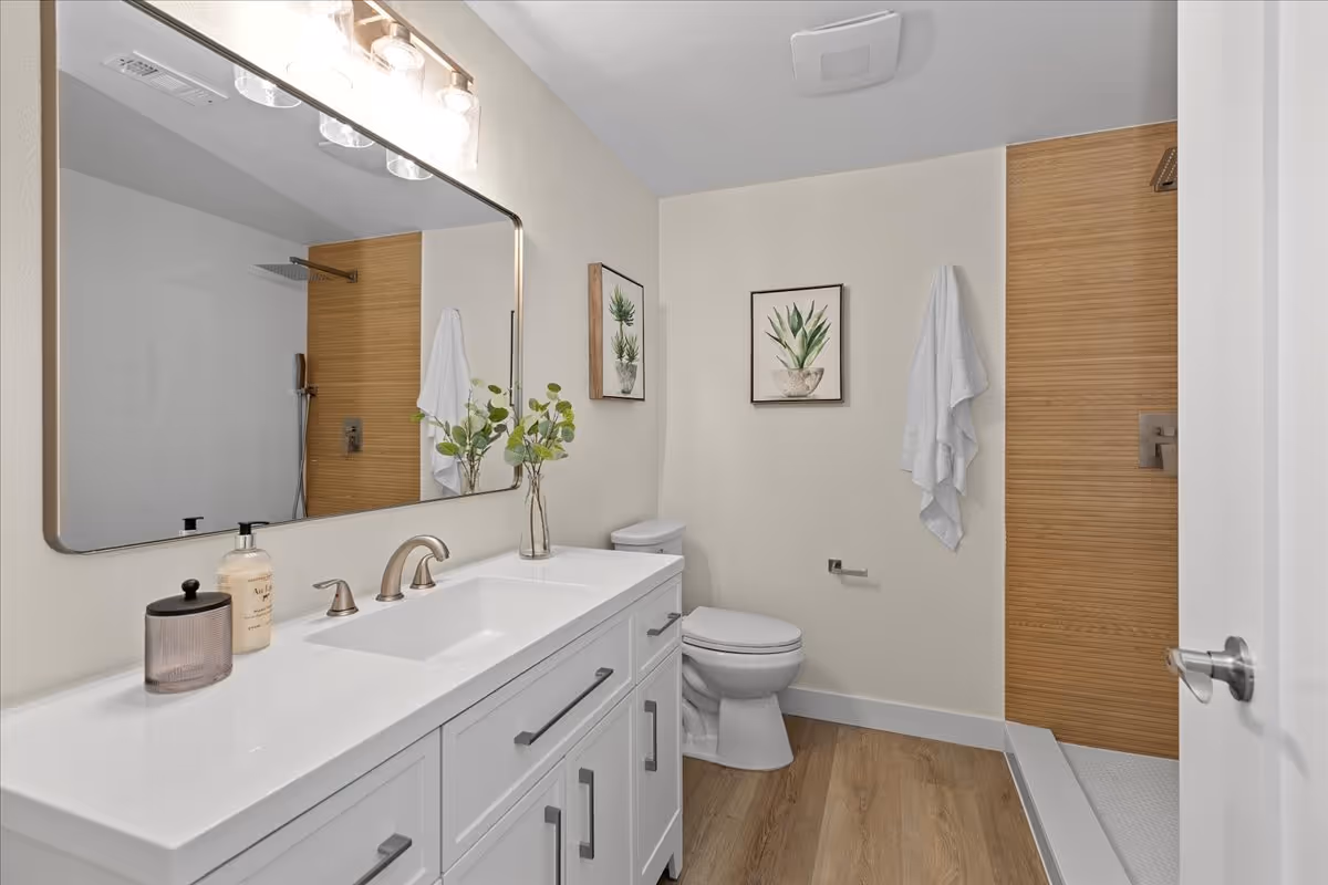 A modern bathroom with a white vanity featuring a sink, faucet, soap dispenser, and a small container. Above the vanity is a large rectangular mirror with three light fixtures. The bathroom has a toilet next to the vanity, two framed plant artworks on the wall, a white towel hanging on a hook, and a walk-in shower with a wooden accent wall and a rainfall showerhead. The floor is wood-style, and the walls are painted white.
