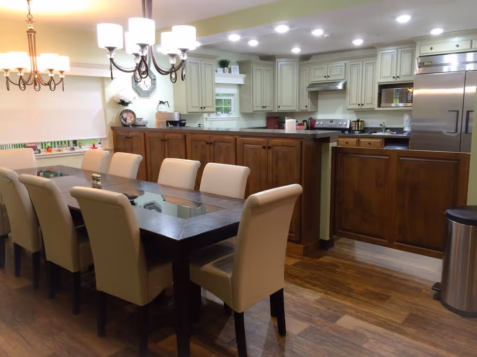 A dining area with a long rectangular glass-top table surrounded by eight beige upholstered chairs. Behind the table is a kitchen with wooden cabinets, a stainless steel refrigerator, stove, microwave, and a countertop with various kitchen items. The room has wooden flooring and is well-lit with ceiling lights and two chandeliers above the dining table.