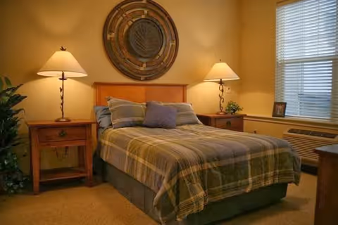 A cozy bedroom with a double bed covered in a plaid comforter and pillows. There are two wooden nightstands on either side of the bed, each with a lamp. A large decorative circular wall piece hangs above the bed. A window with blinds is on the right side, and a small plant and framed photo sit on the windowsill. The room has warm lighting and beige walls.