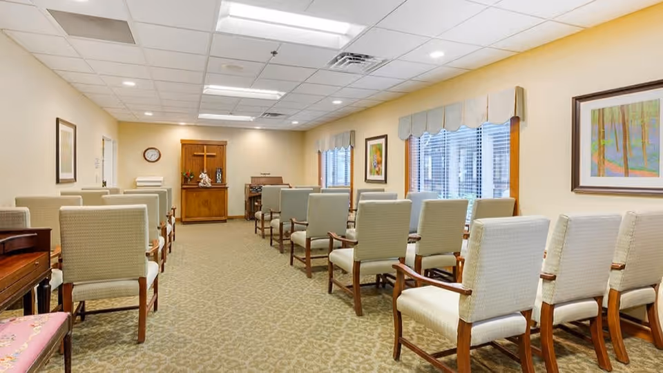 A bright multipurpose meeting room with rows of upholstered chairs facing a wooden cabinet and podium.
