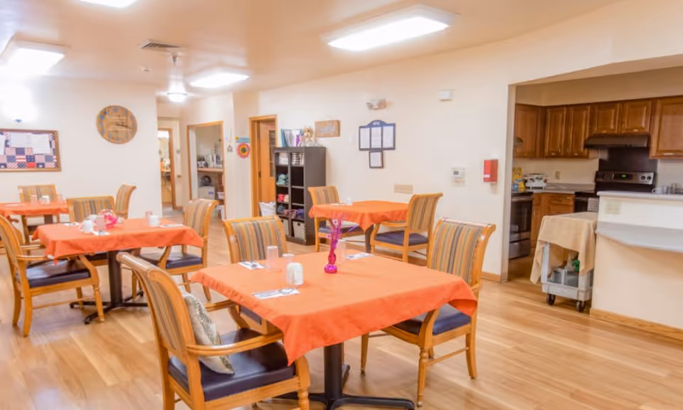 A dining room in an assisted living facility with several tables covered in orange tablecloths, each surrounded by wooden chairs with striped cushions. The room has light-colored walls, wooden flooring, and a kitchen area visible in the background. There are decorations on the walls and a shelving unit with various items.