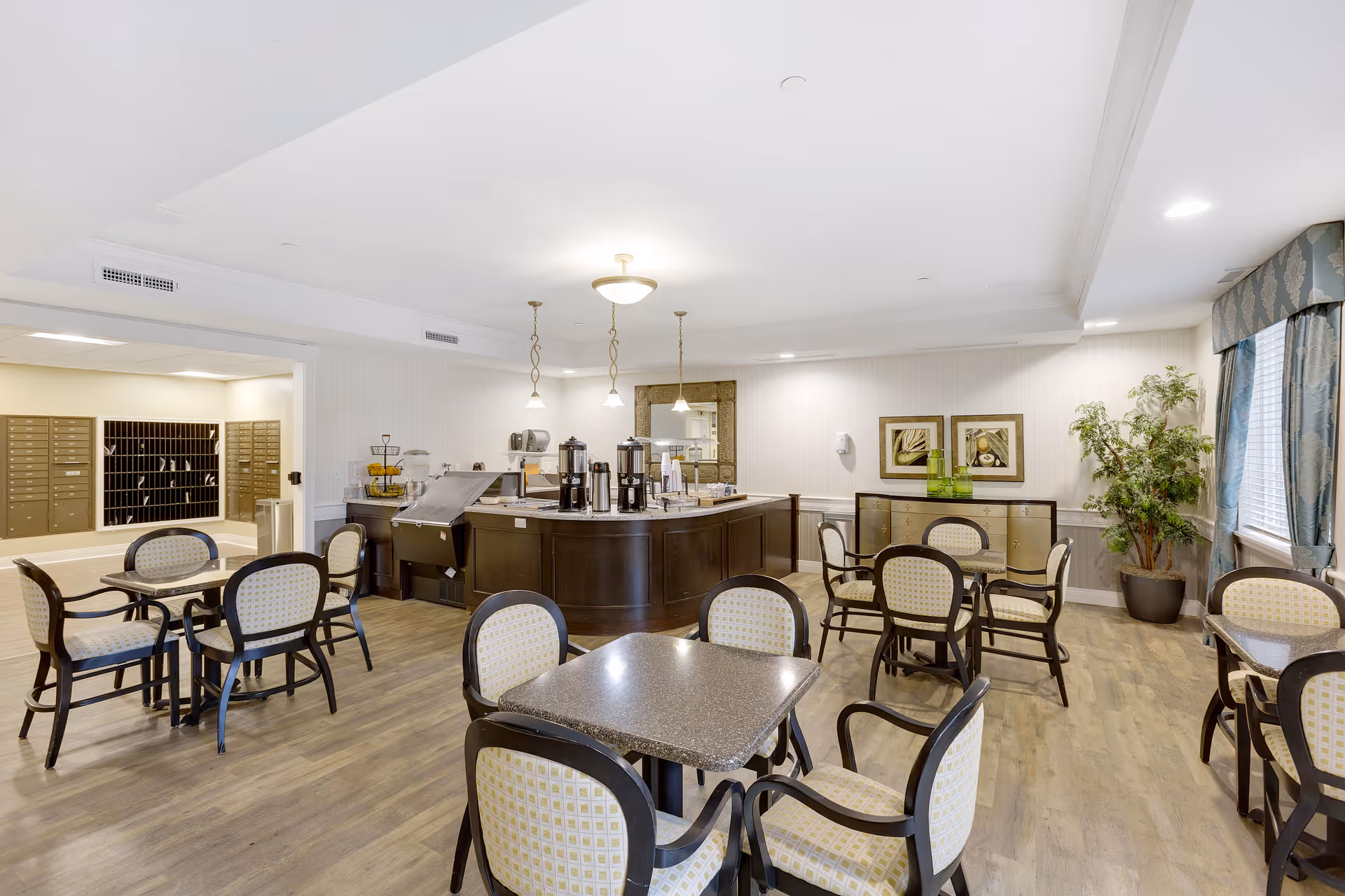 A bright and spacious dining area with multiple tables and chairs arranged neatly. In the center, there is a counter with coffee dispensers and other refreshments. The room has wooden flooring, light-colored walls, framed artwork, a large mirror, and a potted plant near a window with blue curtains. To the left, there is a mail area with multiple mailboxes.