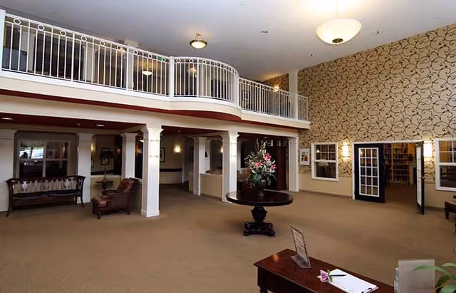 Spacious interior lobby area with beige carpet, a round table with a floral arrangement in the center, white pillars supporting a second-floor balcony with white railing, patterned wallpaper on one wall, and seating areas with chairs and benches.