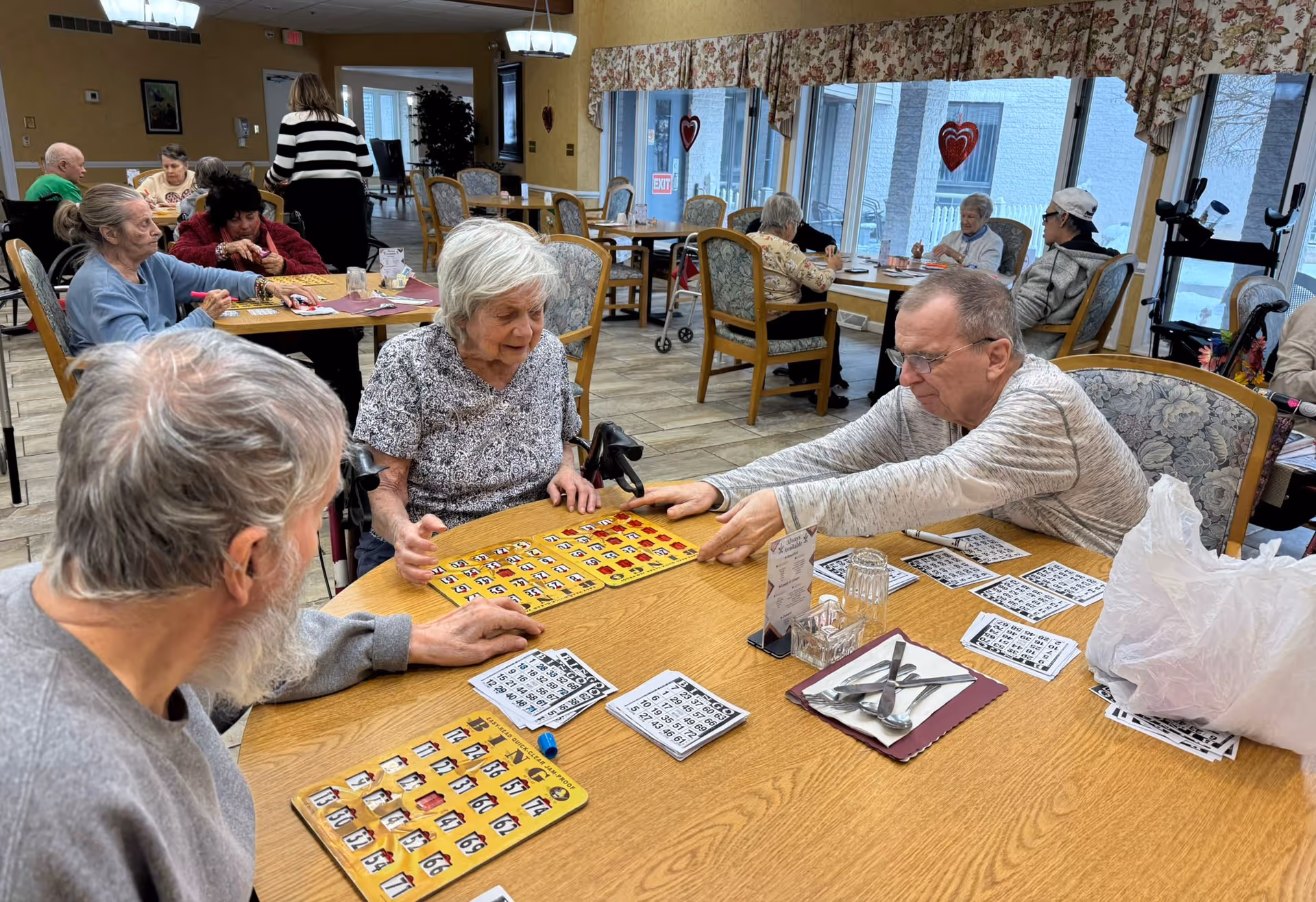 A group of elderly people sitting around tables in a well-lit room playing bingo. The room has large windows with floral curtains and heart decorations. Several other elderly individuals are seated at tables in the background, some engaged in conversation or activities. The atmosphere appears social and communal.