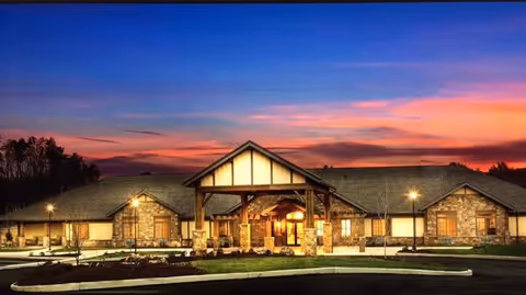 Exterior view of a single-story senior living facility building at sunset with warm lights illuminating the entrance and surrounding area, featuring stone and wood architectural elements and a landscaped front.