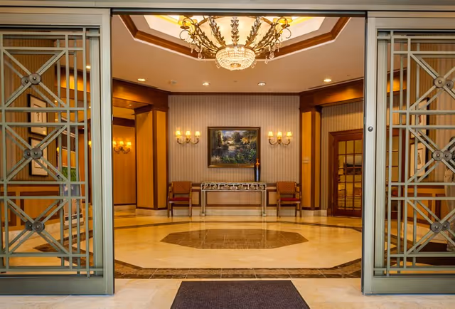 Entrance view into a well-lit, elegant lobby area with decorative metal sliding doors, a chandelier hanging from a recessed ceiling, two chairs flanking a console table with a decorative vase, wall sconces, and a framed landscape painting on the wall.