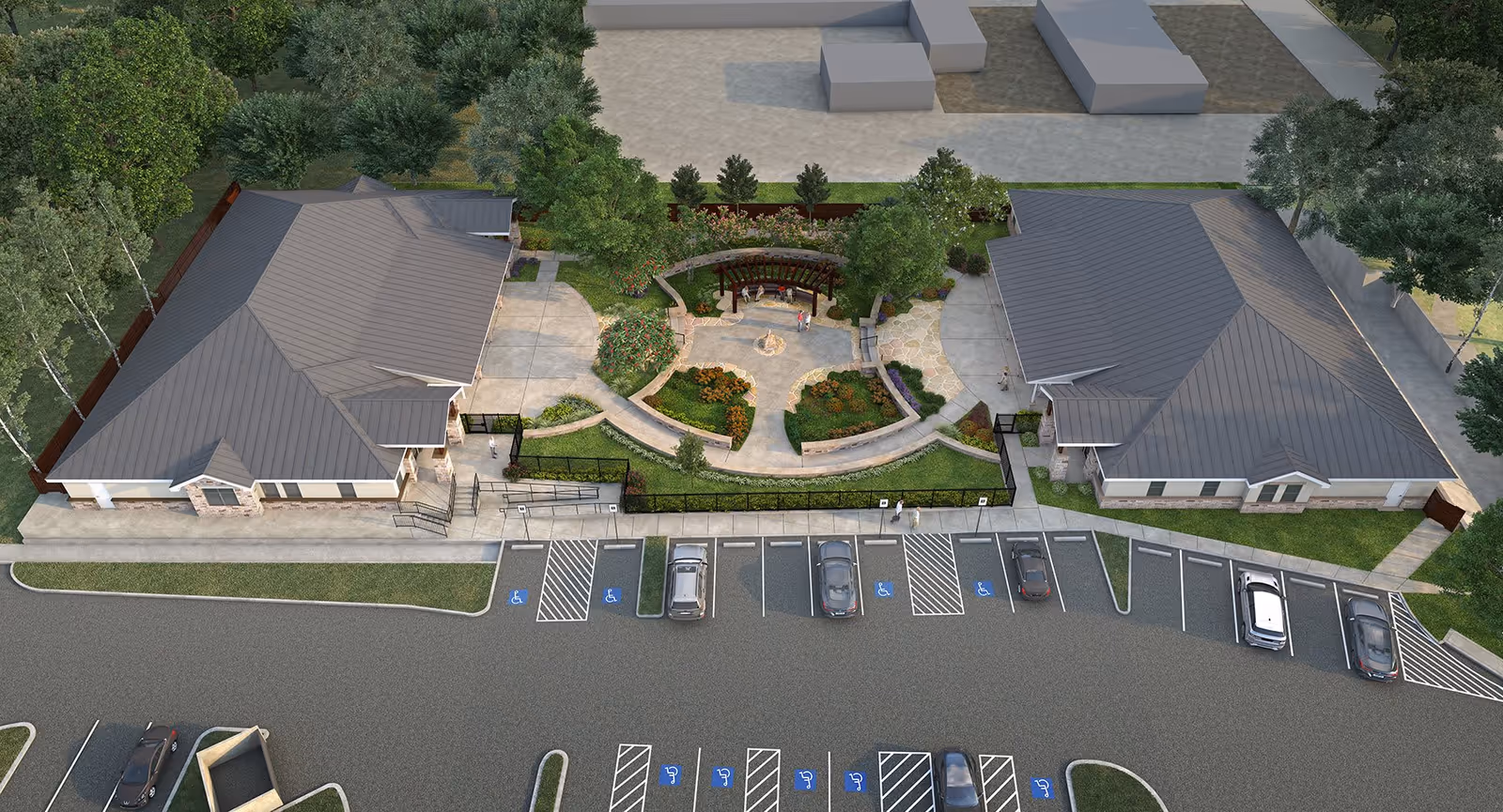 Aerial view of Serenity Oaks Assisted Living and Memory Care facility showing two large buildings with gray roofs separated by a landscaped garden area with pathways, benches, and trees. In front of the buildings is a parking lot with several cars and multiple handicap parking spaces.