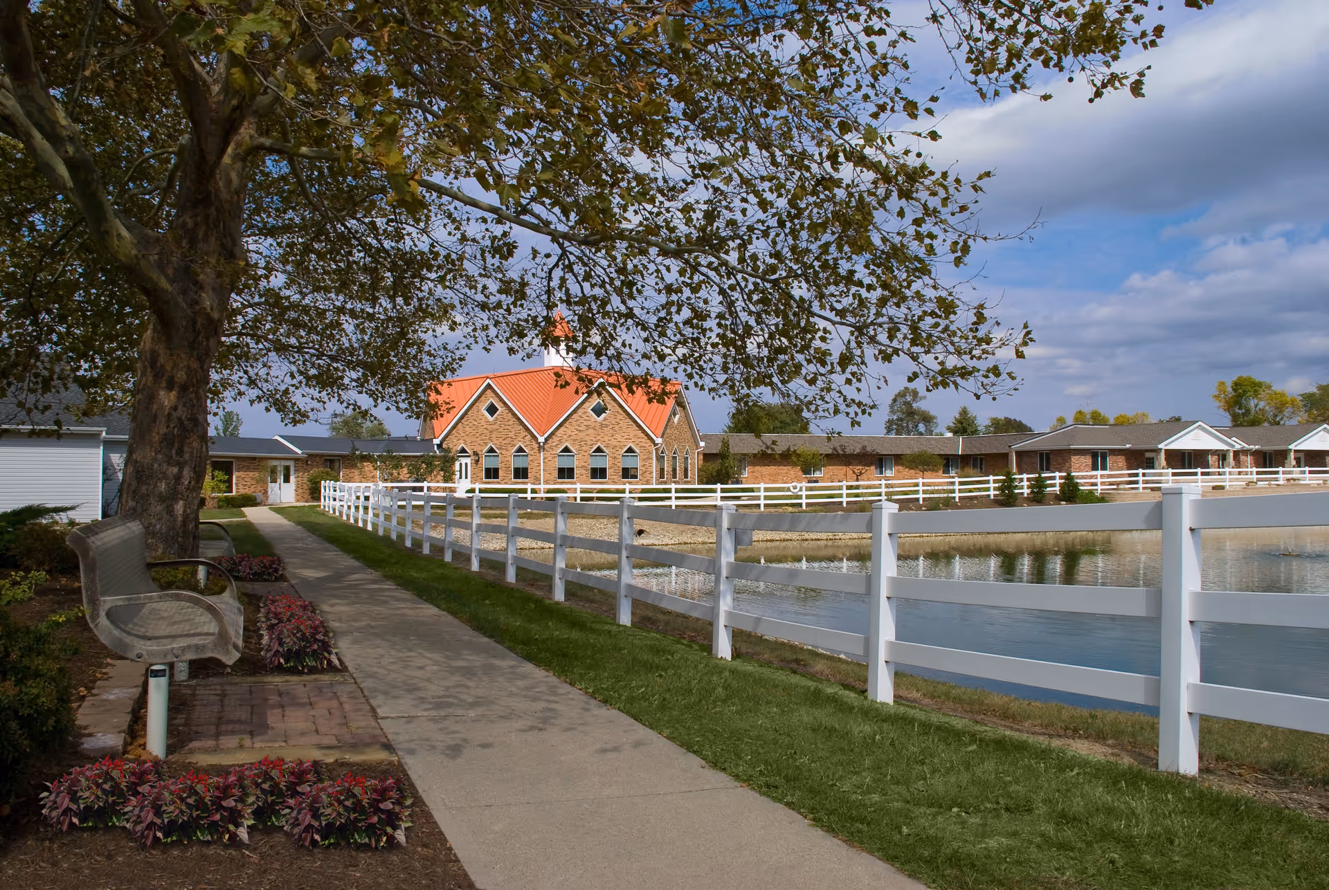 Walkway with benches and a white fence beside a pond leading to a brick building with a red roof beneath a large tree.