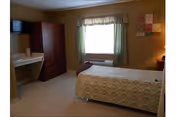 Small nursing-home bedroom with a single bed, window with green curtains, wall-mounted TV, armoire and a small desk.