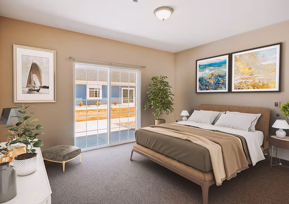 A well-lit bedroom with a large bed covered in beige and brown bedding, two framed abstract paintings above the headboard, a sliding glass door leading outside, a small round side table with a lamp, a potted plant in the corner, a framed picture of a sailboat on the wall, and a cushioned stool near a white dresser with decorative items.