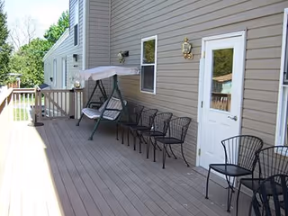 Wooden deck outside a beige-sided building with a canopy swing, several metal chairs, and a white door.