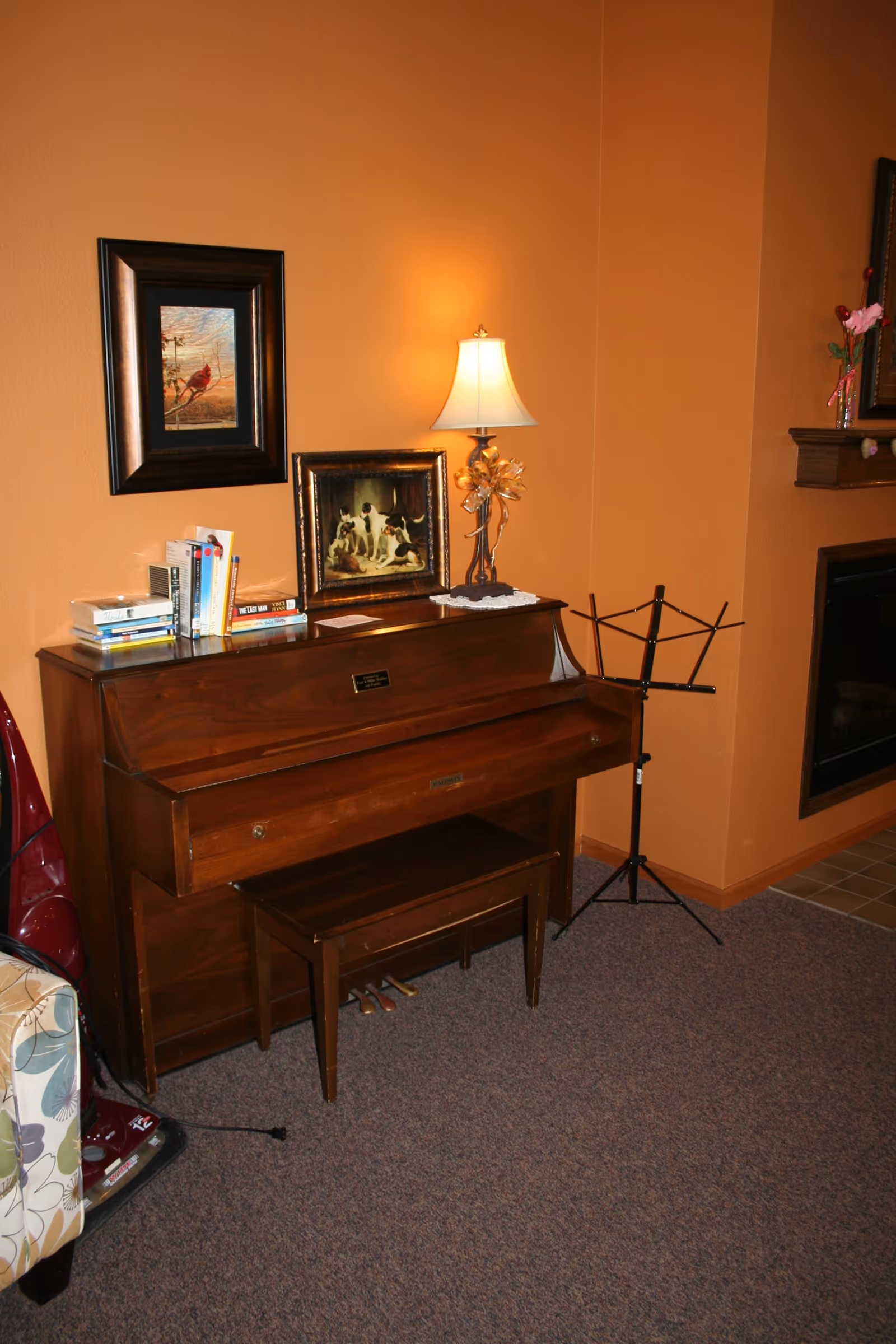 A cozy corner of a living room with an upright wooden piano and matching bench. On top of the piano are several books, a framed painting of dogs, and a table lamp with a decorative base. A music stand is positioned next to the piano. The walls are painted a warm orange color, and part of a fireplace and a floral-patterned armchair are visible.