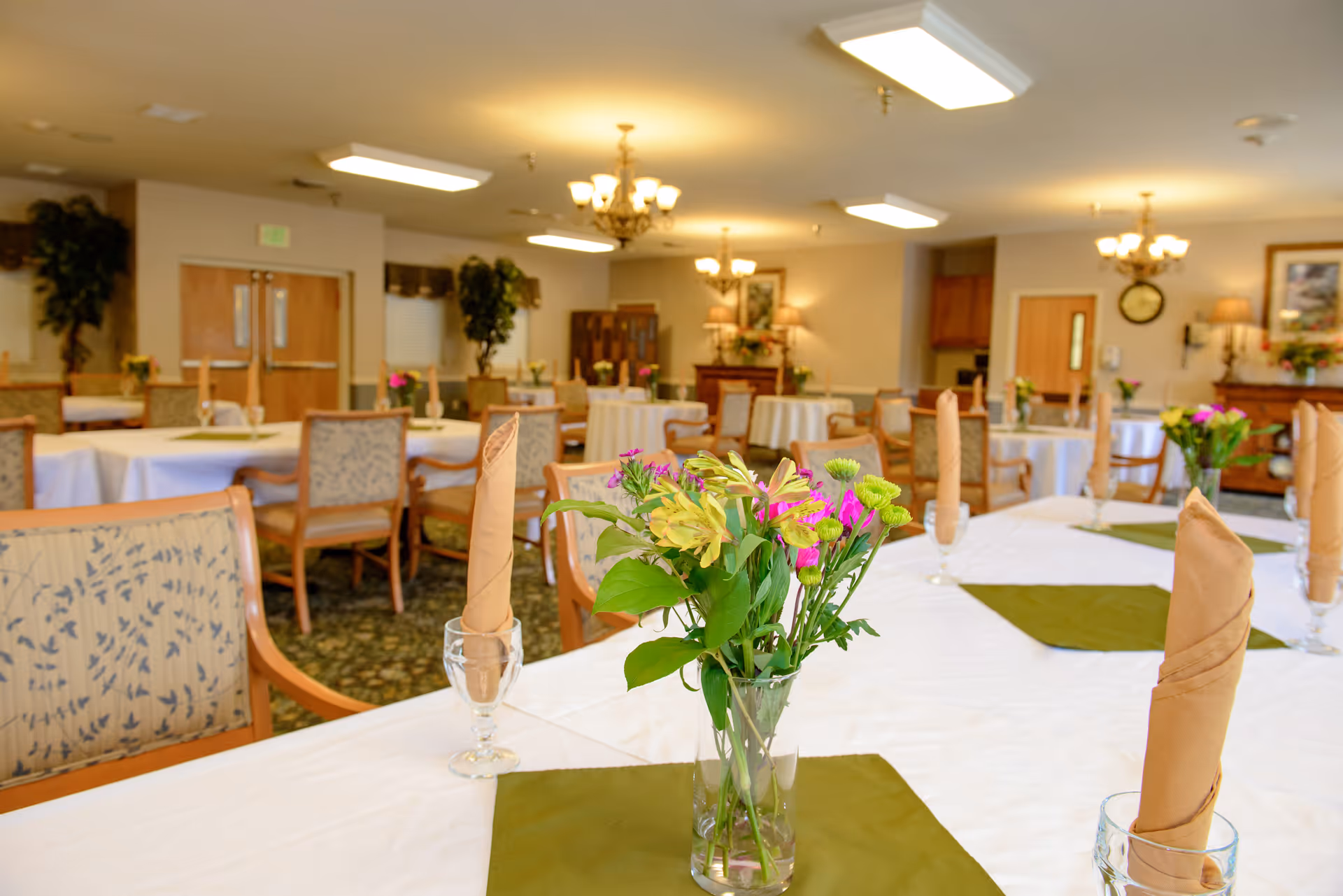 A well-lit dining room in a senior living facility with multiple round tables covered in white tablecloths and green placemats. Each table has a centerpiece of fresh flowers in a glass vase and neatly folded beige napkins standing upright in glass holders. The room features wooden chairs with patterned upholstery, chandeliers, and decorative plants and furniture along the walls.