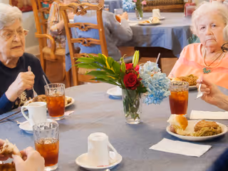 Elderly individuals seated around a dining table with plates of food and glasses of iced tea, with a floral centerpiece on the table in a communal dining setting.