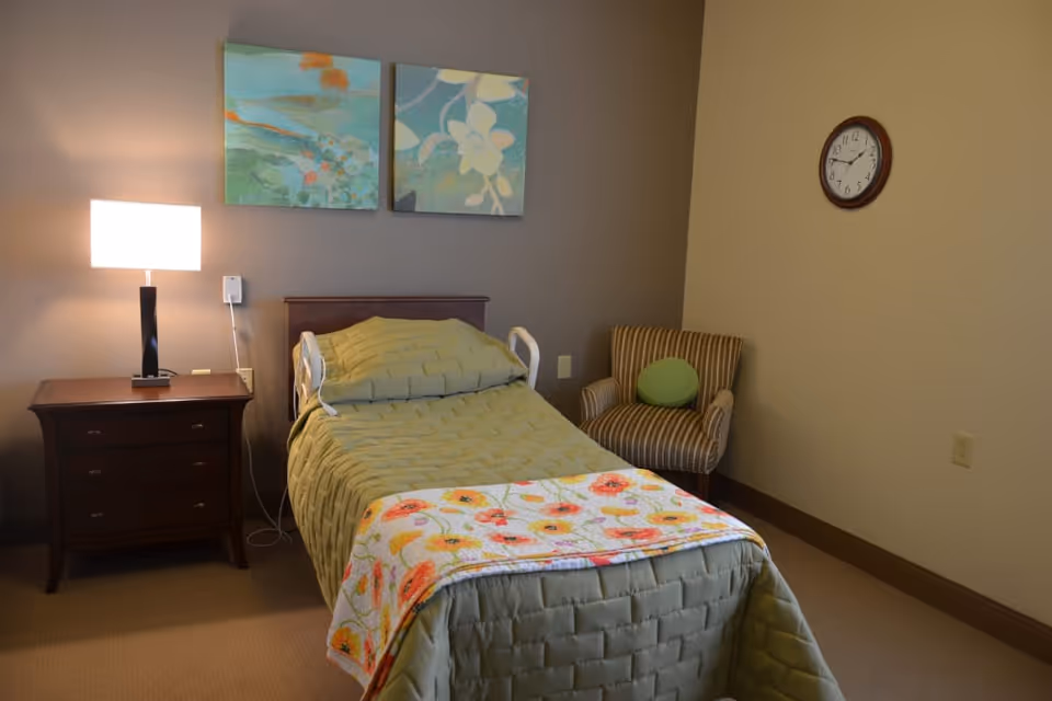A single bed with a green quilt and a floral blanket at the foot is positioned against a beige wall. To the left of the bed is a wooden nightstand with a modern lamp emitting warm light. Above the bed are two abstract paintings in shades of green and blue. To the right of the bed is a striped armchair with a green round pillow. A round wall clock is mounted on the right wall.