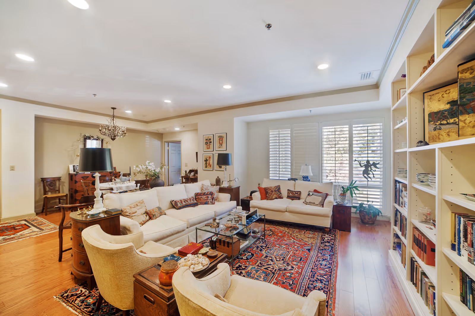 A spacious and well-lit living room featuring two white sofas with decorative pillows, two beige armchairs, a glass coffee table, and a large colorful patterned rug. The room has wooden flooring, built-in bookshelves filled with books and decor items, large windows with white shutters, and various lamps and artwork on the walls.