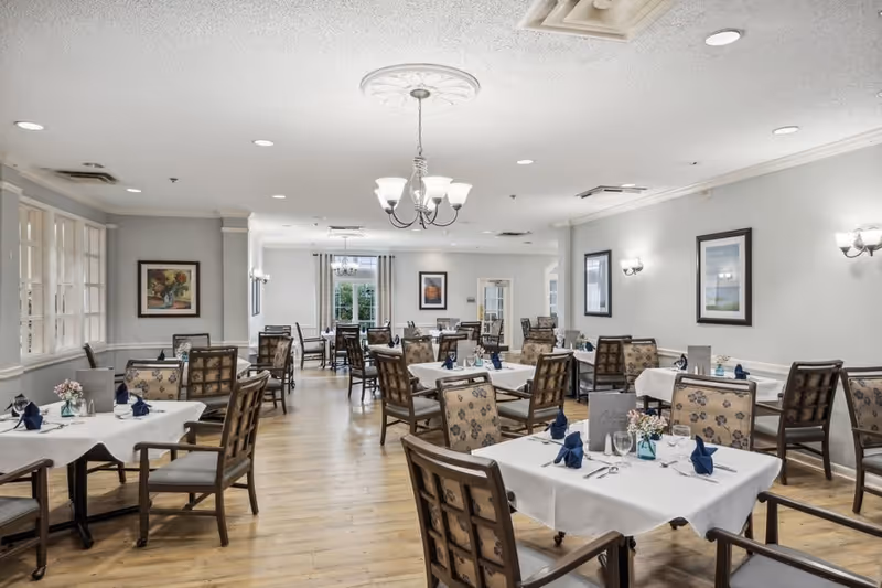 A spacious dining room with multiple tables covered in white tablecloths, each set with blue napkins, glassware, and silverware. The room has wooden flooring, light gray walls adorned with framed artwork, and several chandeliers and wall sconces providing lighting. Large windows on one side allow natural light to enter.